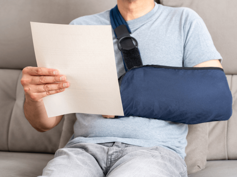 Man with arm in sling reads document while sitting on sofa.