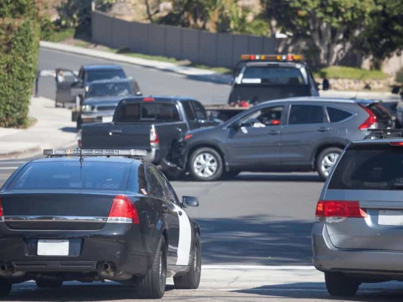 A black sedan with a light bar is parked on a street, observing a car accident on the highway in a suburban setting.