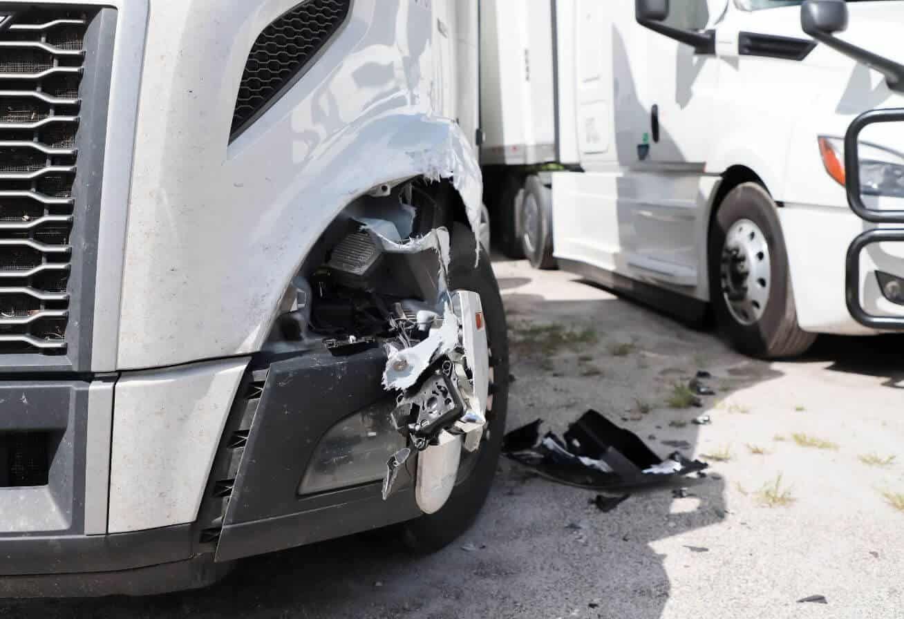 A damaged white truck with a destroyed front bumper, parked on an asphalt surface with debris scattered around, suggesting a recent collision.