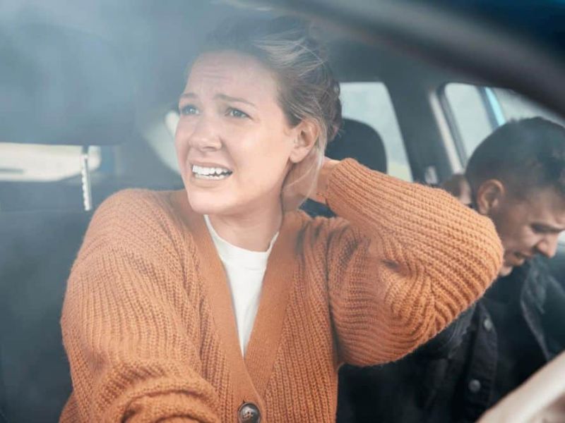 A concerned woman braces herself in a car, focusing on something outside, while a man appears to be searching for something in the front seat, creating a tense atmosphere.