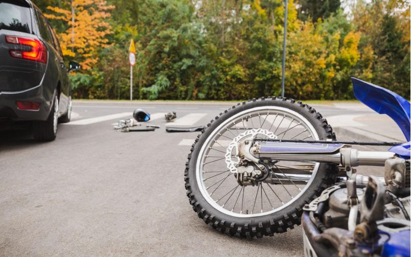 Car and motorcycle accident at intersection with scattered parts and fall foliage background.