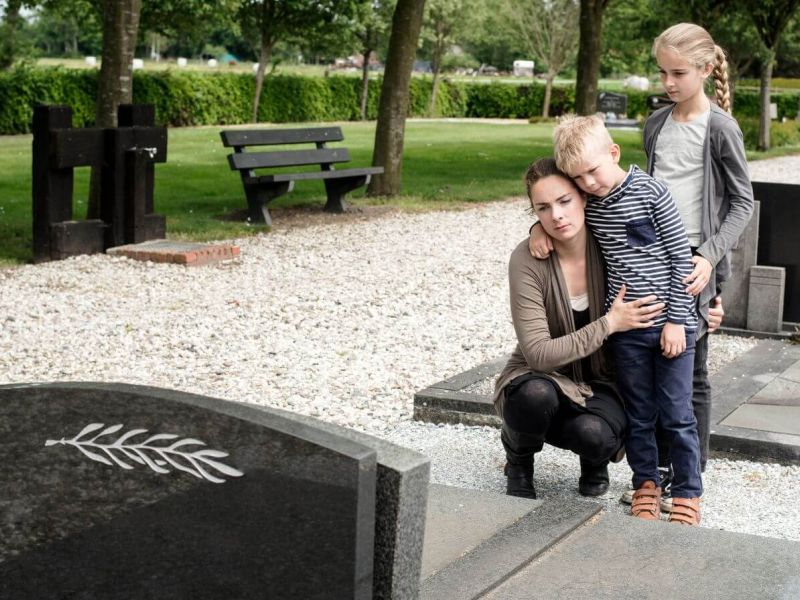 A woman crouches beside a gravestone, comforting a young boy who leans on her, while an older girl stands nearby, all in a serene cemetery surrounded by green trees and benches.