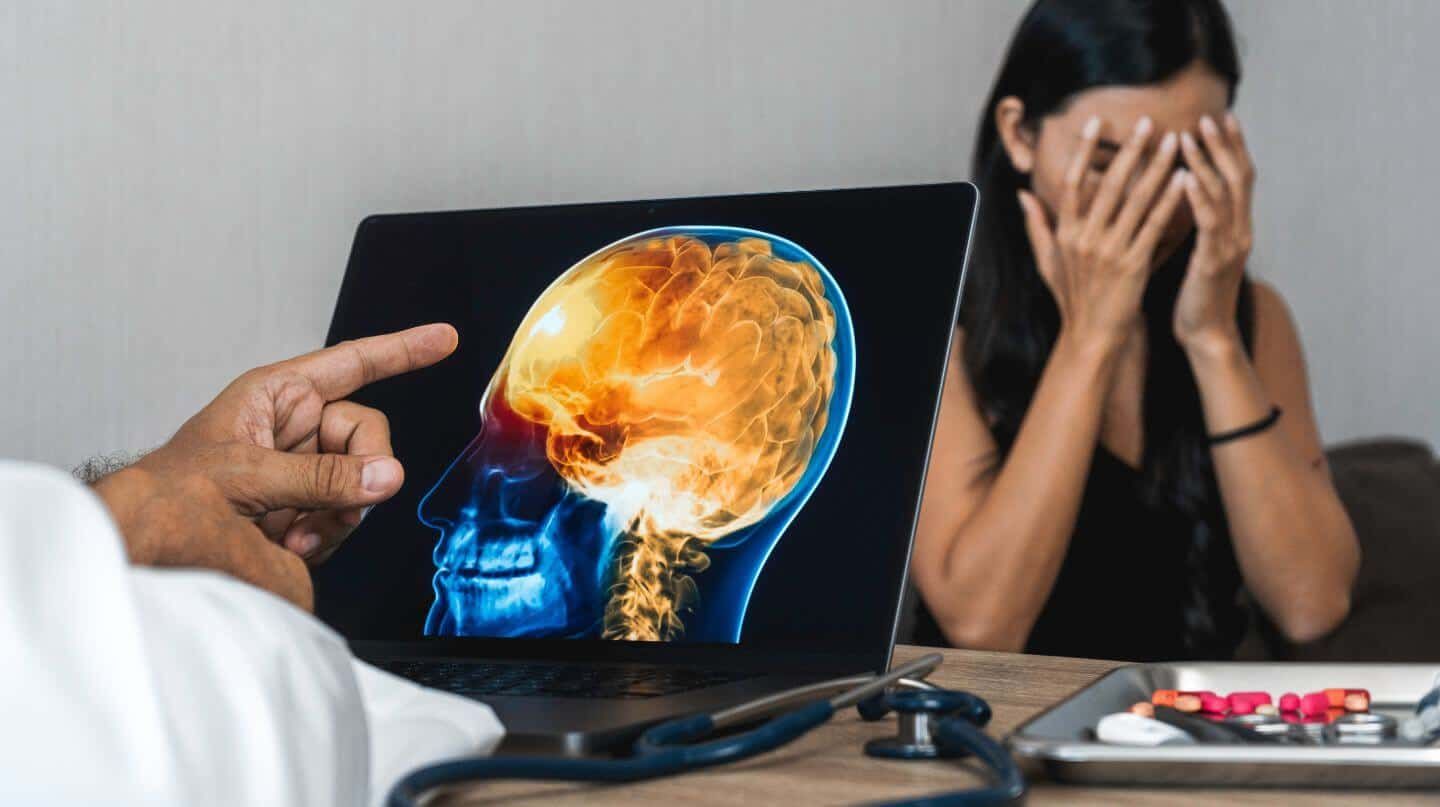 A doctor points to a brain MRI on a laptop screen, illustrating a problem, with a distressed woman in the background; a stethoscope and pills are on the table.