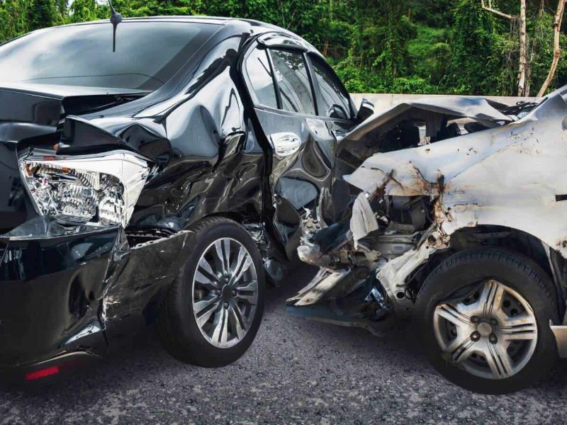 Two cars with frontal damage after a collision, amid a road setting with green foliage in the background.