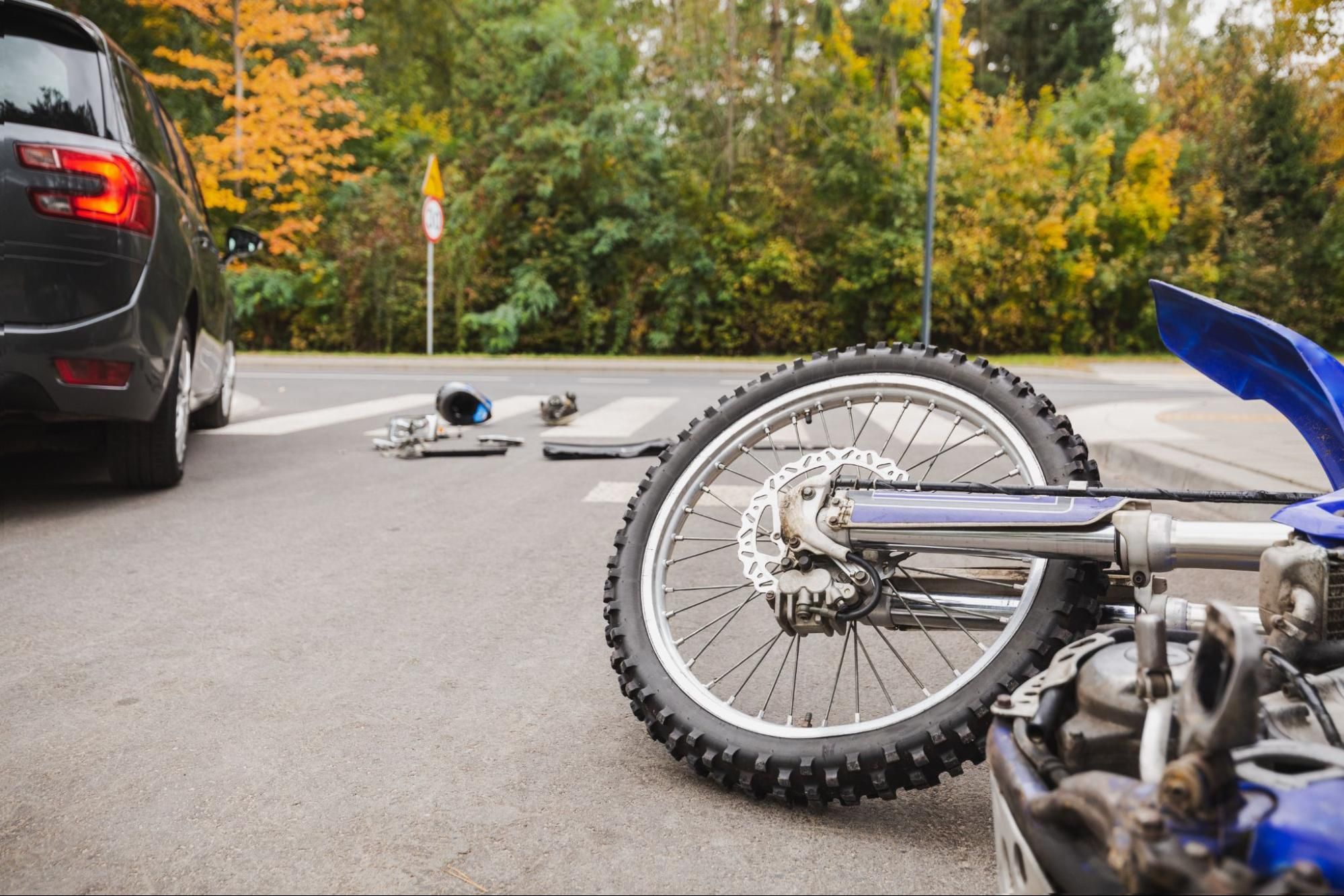 Car and motorcycle accident at intersection with scattered parts and fall foliage background.