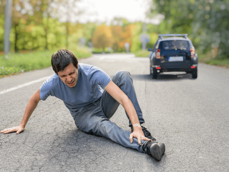A man is sitting on the road, grimacing in pain while holding his ankle. A black car approaches in the background, surrounded by green trees and a clear sky.