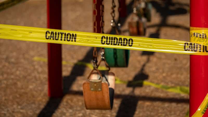 Yellow caution tape with "CAUTION CUIDADO" spans across the frame, obstructing access to a blurred playground with swing sets in the background.