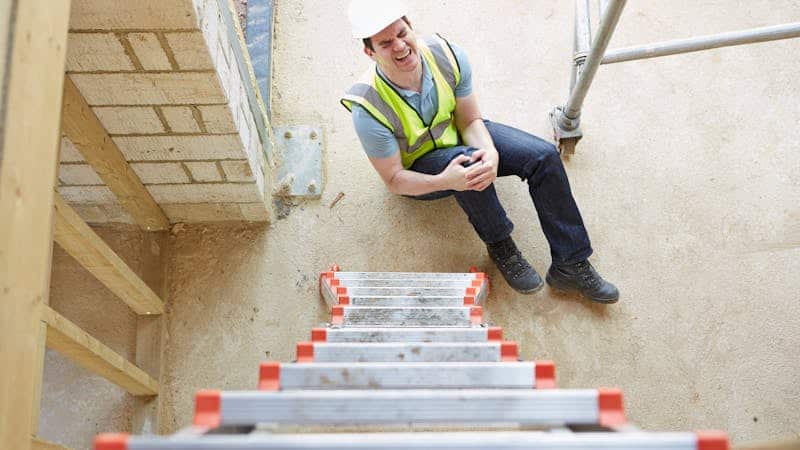 A construction worker in a hard hat and hi-vis vest sits injured on scaffolding steps, grasping his knee, amidst a construction site.
