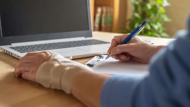 A person with a wrist brace is writing on a clipboard next to a laptop, suggesting work or study in a home or office setting.
