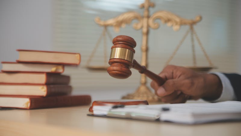 A gavel is raised above a desk where law books are stacked. In the background, a scale of justice symbolizes legal proceedings, creating a courtroom context.