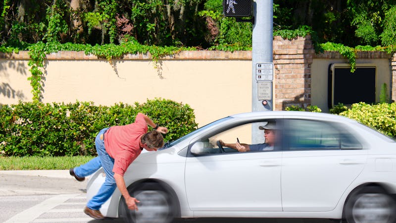 A man in a red shirt is hit by a speeding white car at a crosswalk while the driver appears distracted. The background features a brick wall and greenery.