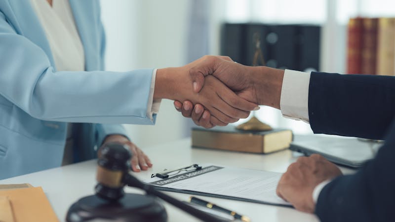 A man and a woman are shaking hands across a table, with a legal document and a gavel visible in the context of a professional office setting.