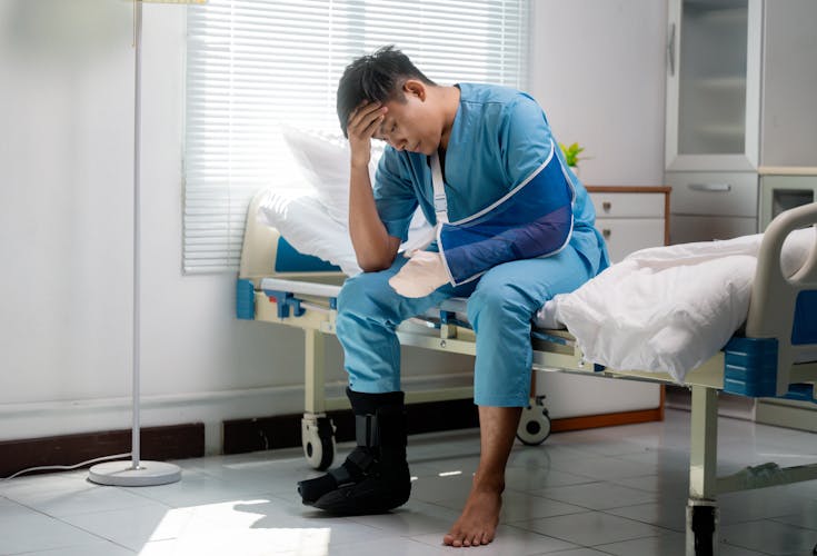 Injured man in hospital gown, wearing arm sling and leg brace, sitting on hospital bed looking distressed.